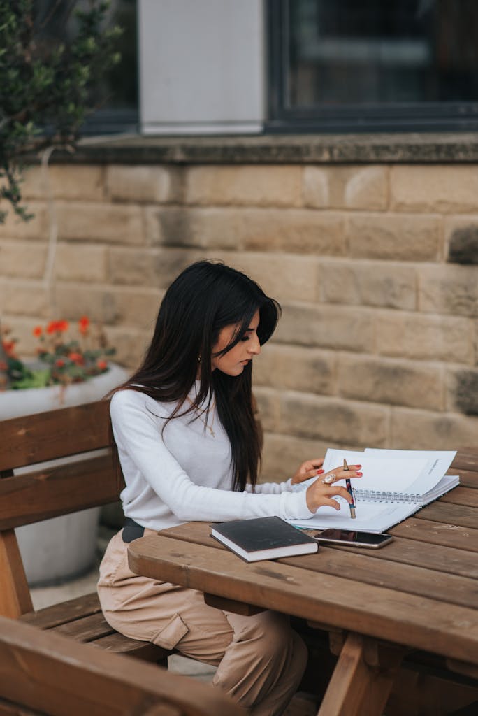 Young woman concentrating on her writing at an outdoor table against a brick wall backdrop.